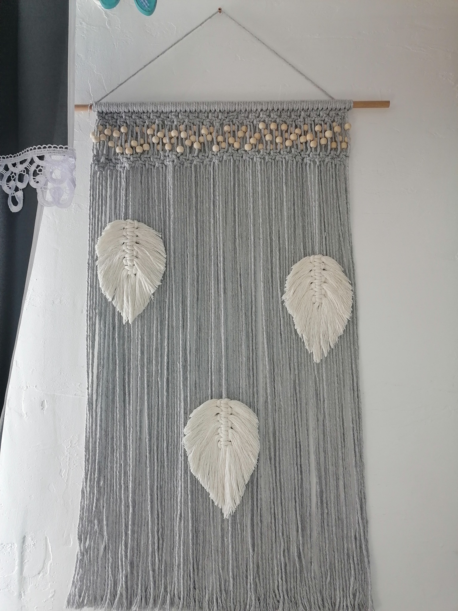 Szara makrama ścienna z trzema białymi piórami, zawieszona na drewnianym drążku z ozdobnymi drewnianymi kulkami.