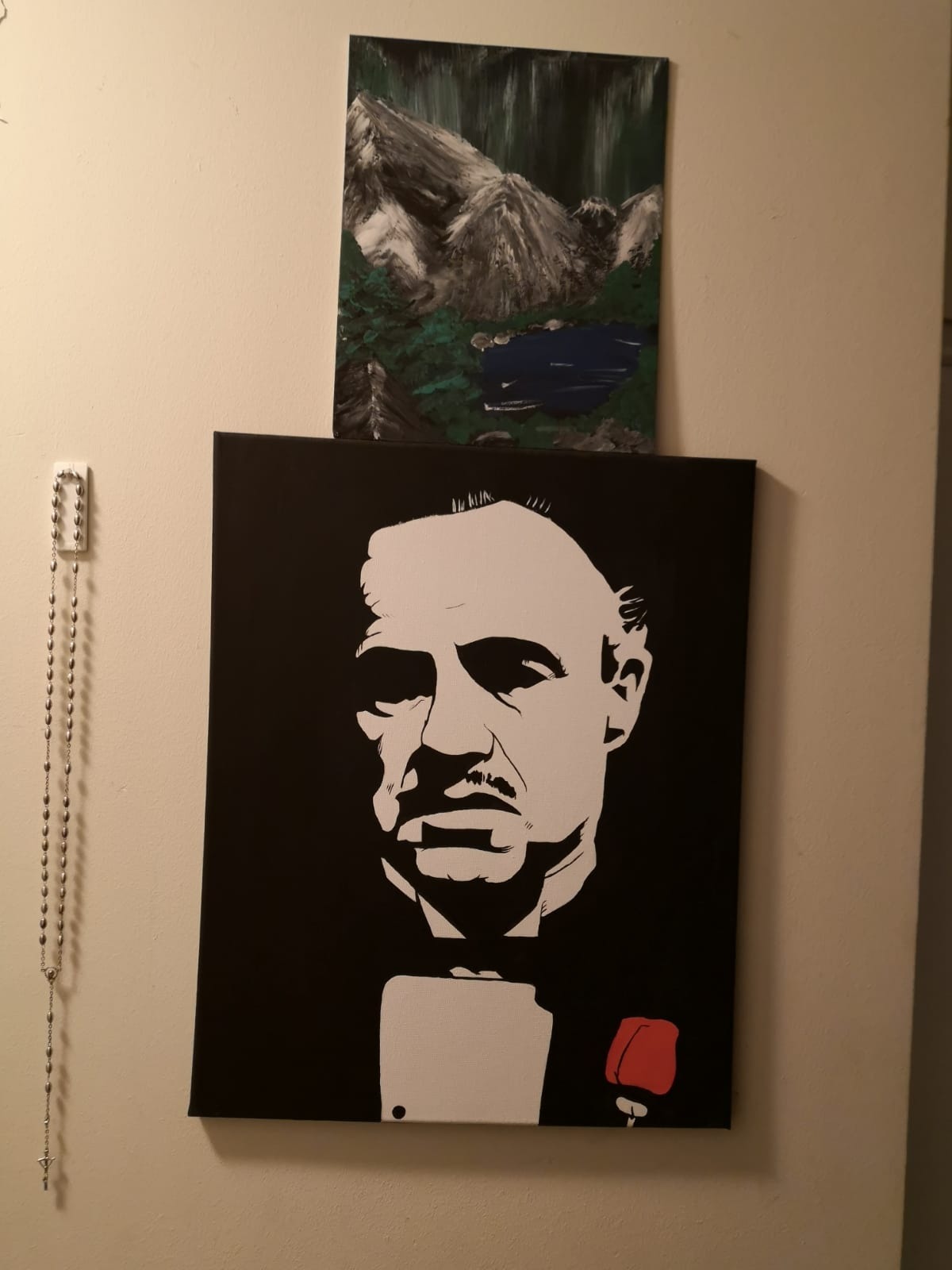 Dwa obrazy na ścianie: na dole czarno-biały portret mężczyzny z czerwoną różą, u góry abstrakcyjny pejzaż górski z jeziorem, malowane farbami.