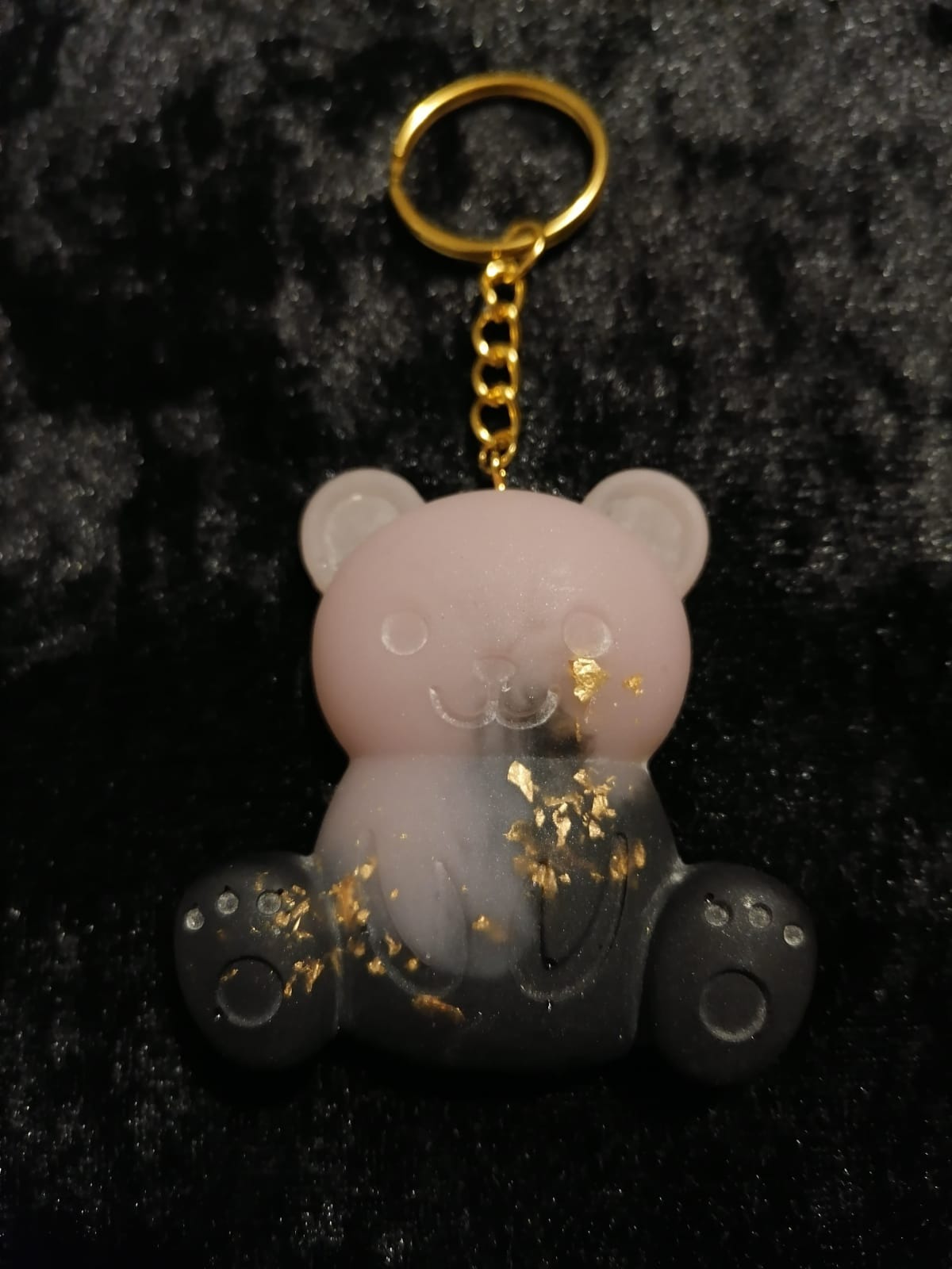 Ręcznie robiony brelok w kształcie misia z żywicy epoksydowej, z dodatkiem złotych płatków, wykonany w Kołobrzegu. Górna część misia jest różowa, a dolna czarna, całość zawieszona na złotym kółku.