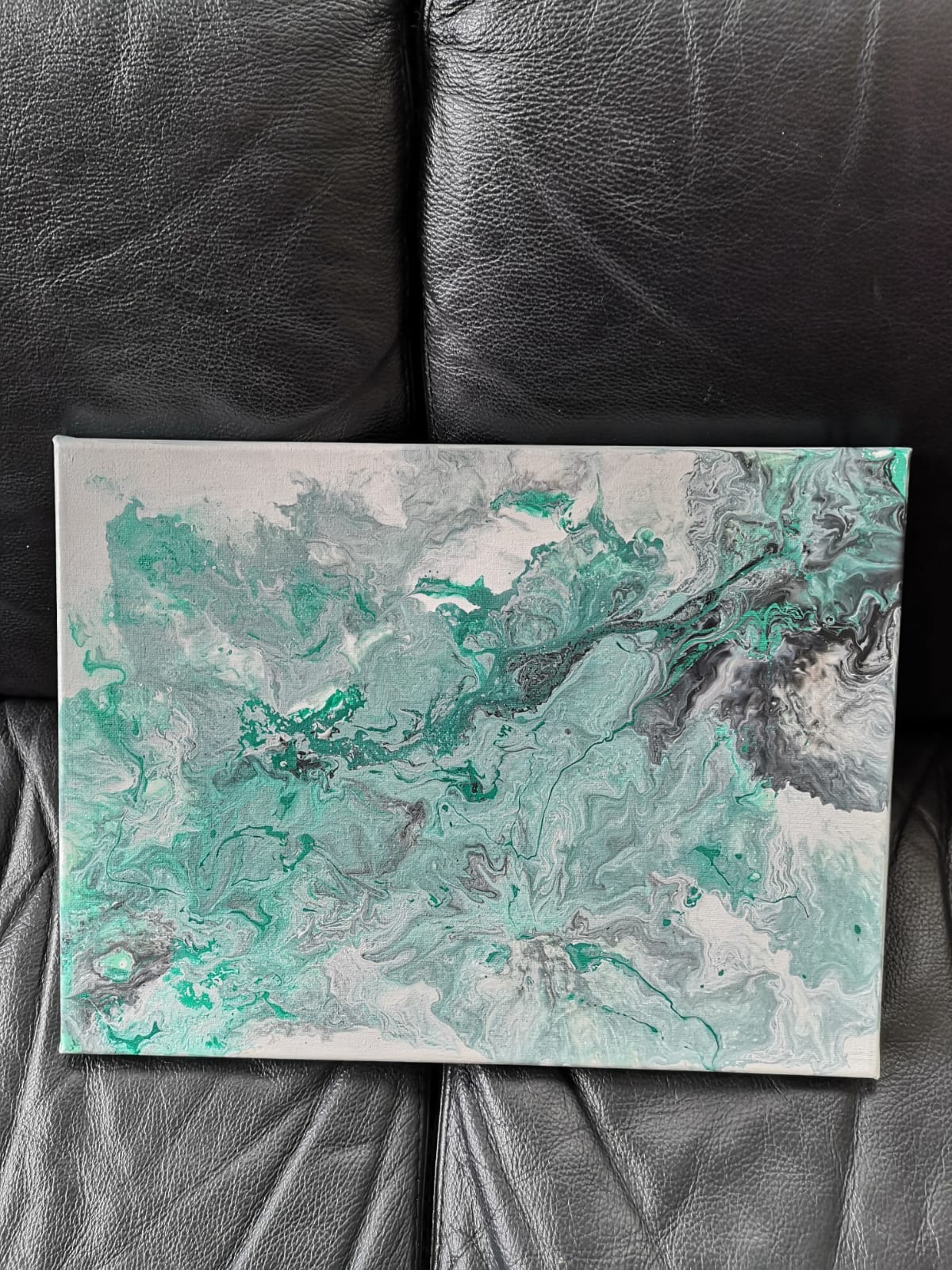 Abstrakcyjne malowidło akrylowe w odcieniach bieli, szarości, czerni i zieleni, wykonane techniką 'pouring' na płótnie, leżące na czarnej, skórzanej powierzchni.