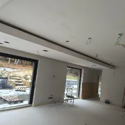 Wnętrze w trakcie prac wykończeniowych we Wrocławiu: sufit z zamontowaną płytą karton gips, widoczne otwory na oświetlenie, ściany przygotowane pod malowanie, duże okna z widokiem na zimowy krajobraz.