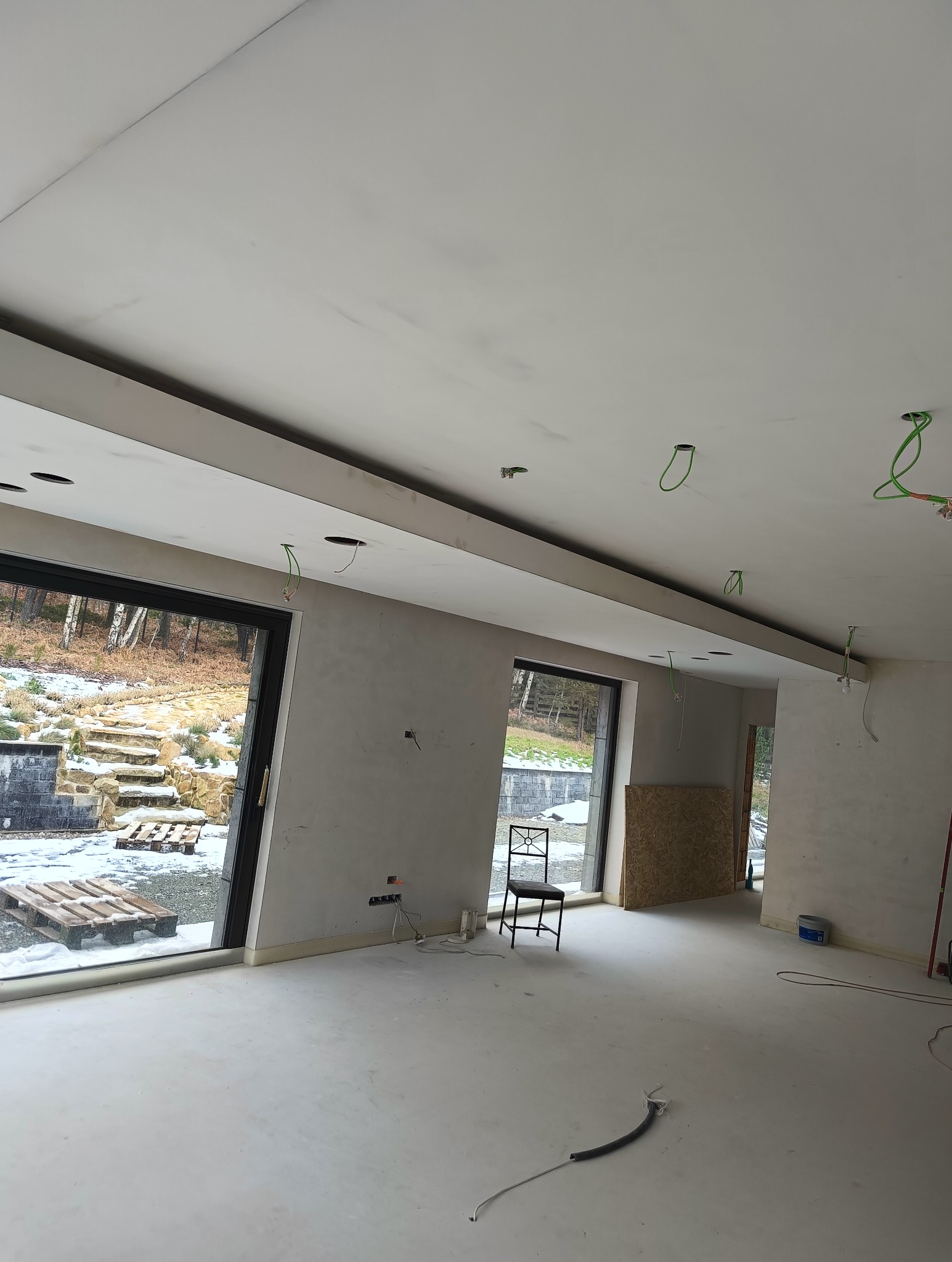 Wnętrze w trakcie prac wykończeniowych we Wrocławiu: sufit z zamontowaną płytą karton gips, widoczne otwory na oświetlenie, ściany przygotowane pod malowanie, duże okna z widokiem na zimowy krajobraz.