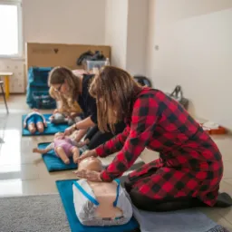 Uczestnicy szkolenia ćwiczą resuscytację krążeniowo-oddechową na manekinach dziecięcych i niemowlęcych, ustawionych na niebieskich matach, w jasnym pomieszczeniu.