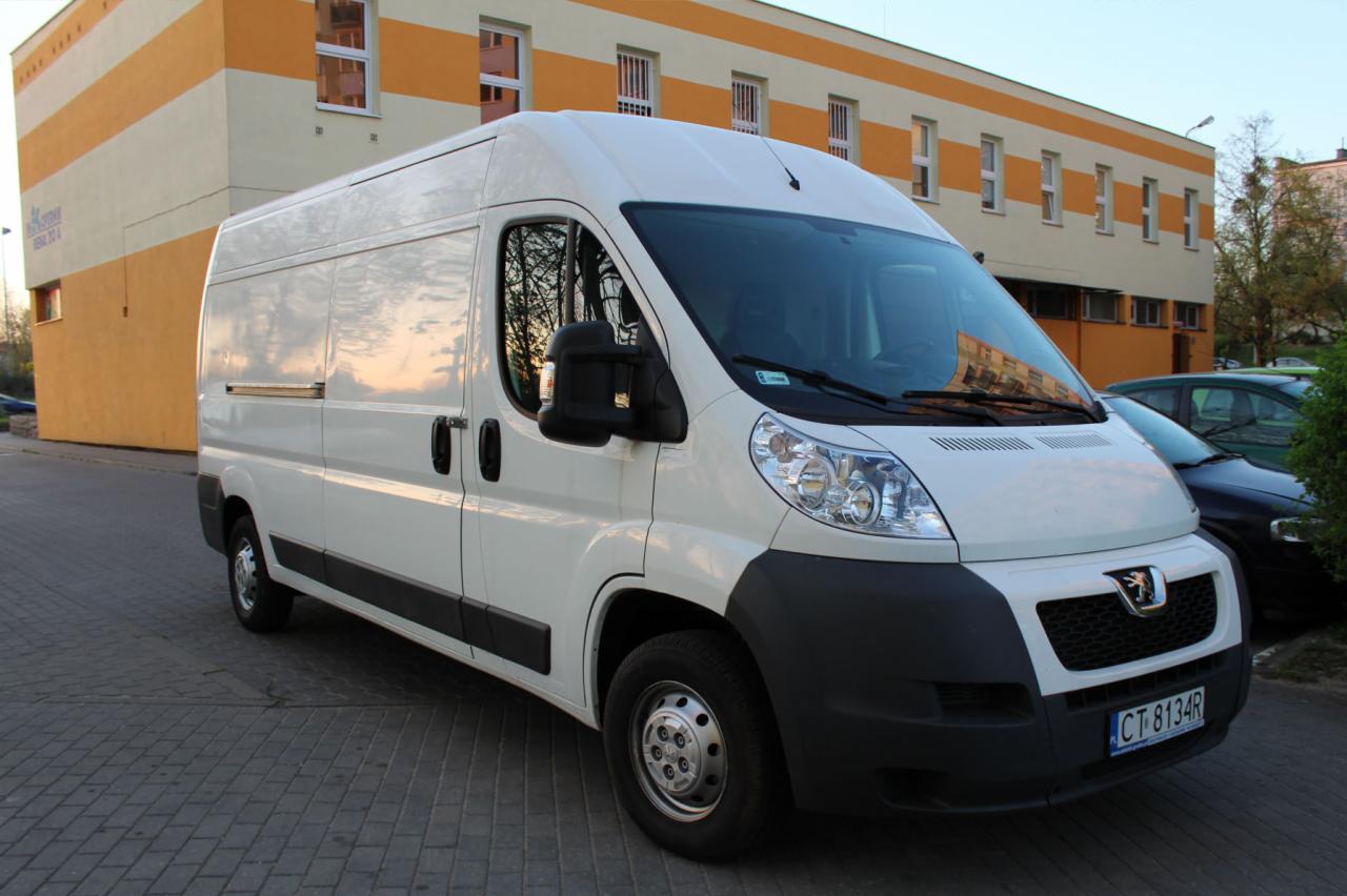 Biały bus marki Peugeot zaparkowany na brukowanej nawierzchni przed budynkiem z pomarańczowo-białymi pasami, widoczny numer rejestracyjny.