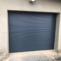 Zewnętrzna roleta w kolorze grafitowym, zamontowana w betonowej wnęce garażowej, widoczna z bliska, z fragmentem brukowanej nawierzchni u dołu.