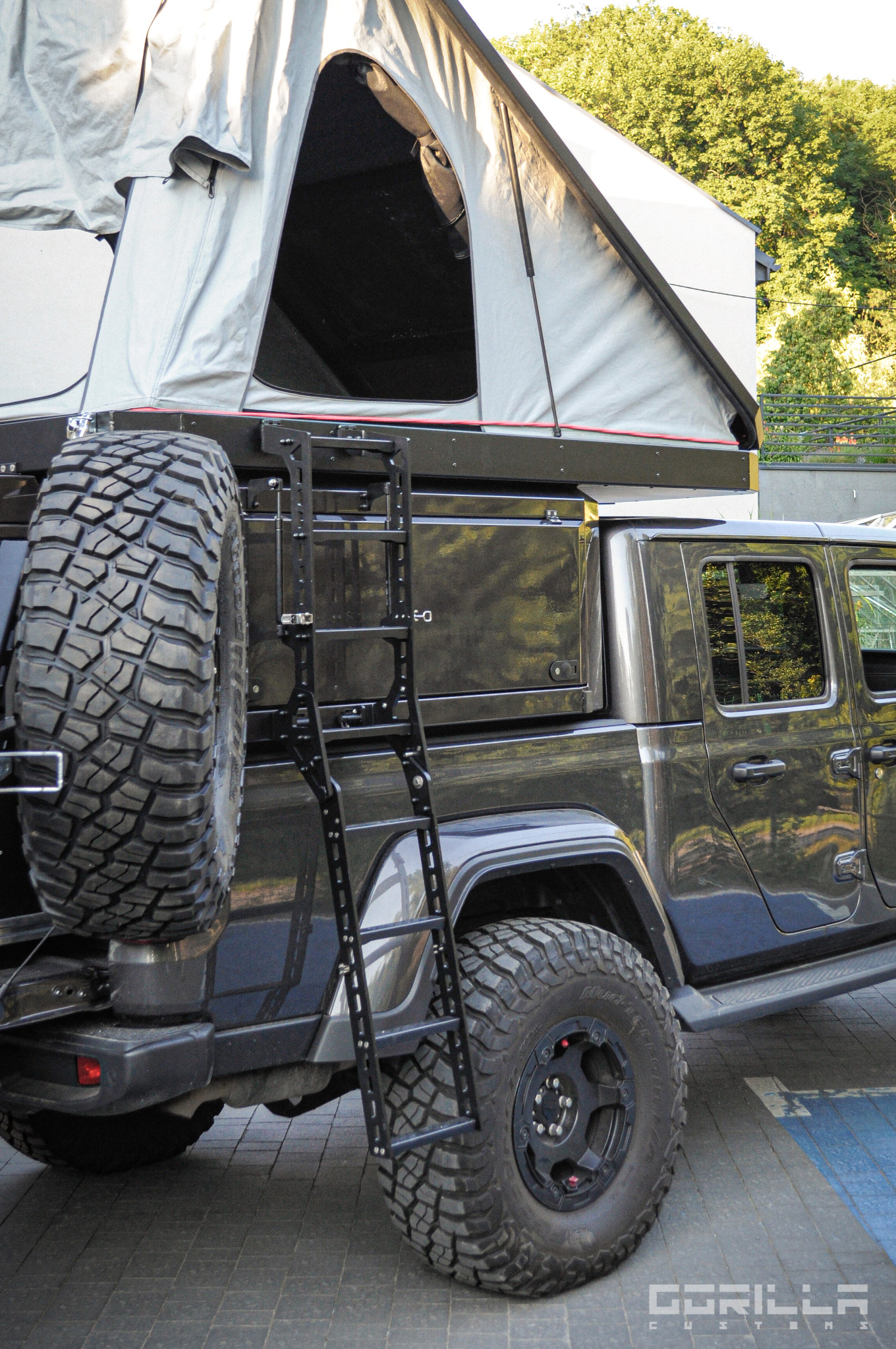 Szary Jeep Gladiator z zamontowanym namiotem dachowym, drabinką i kołem zapasowym na tylnej klapie.