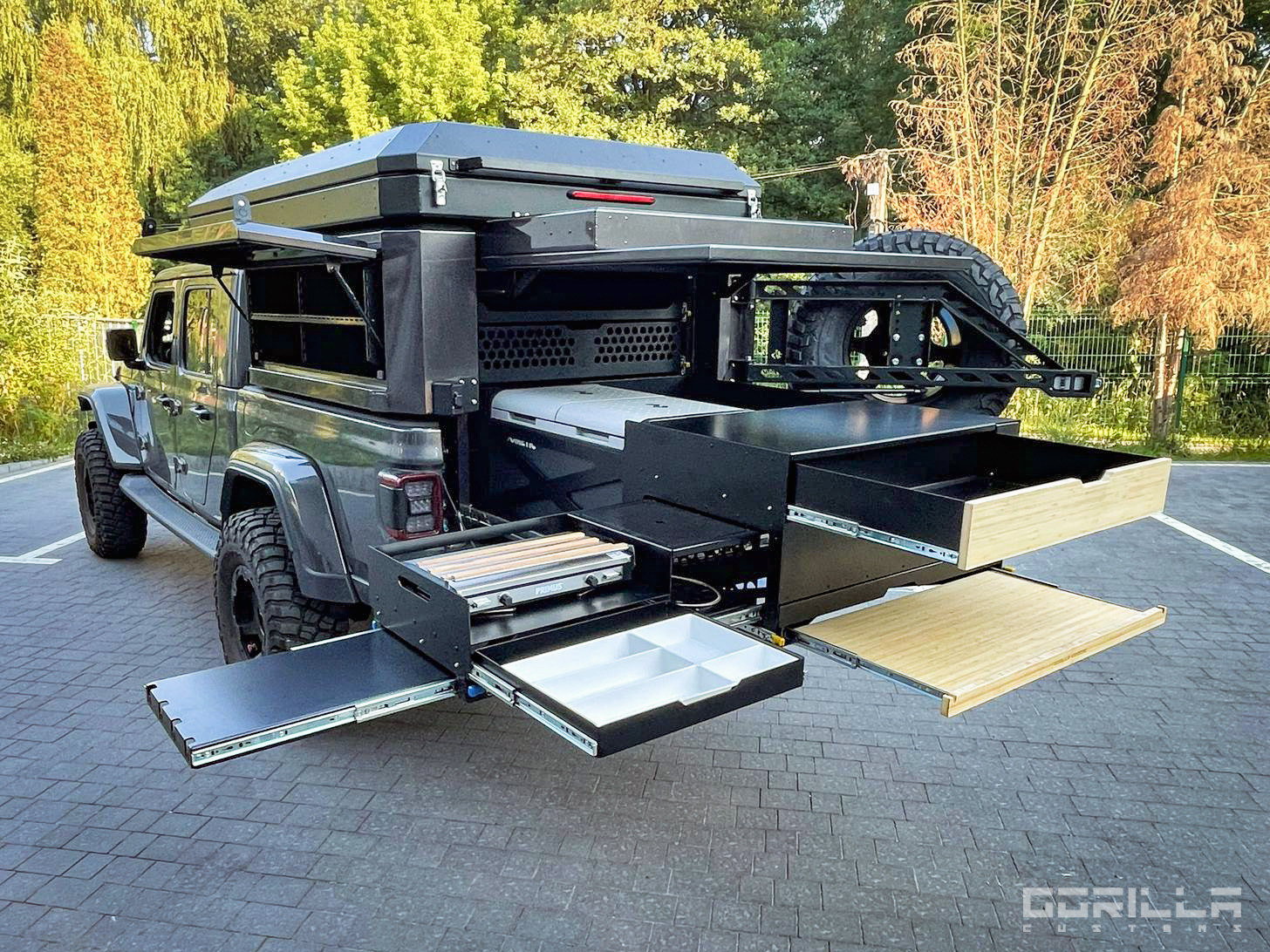 Szary samochód terenowy z zamontowaną na pace zabudową kempingową z wysuwanymi szufladami, blatem i kuchenką gazową, widok z tyłu na tle zieleni.