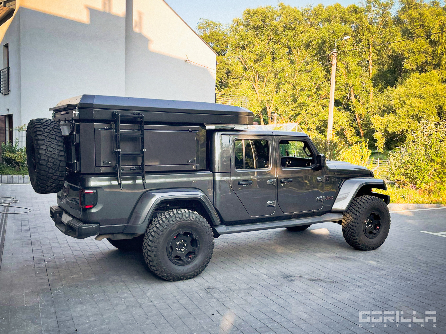 Szary Jeep Gladiator z zabudową kempingową i drabinką, zaparkowany w Krakowie na tle budynku i zieleni.