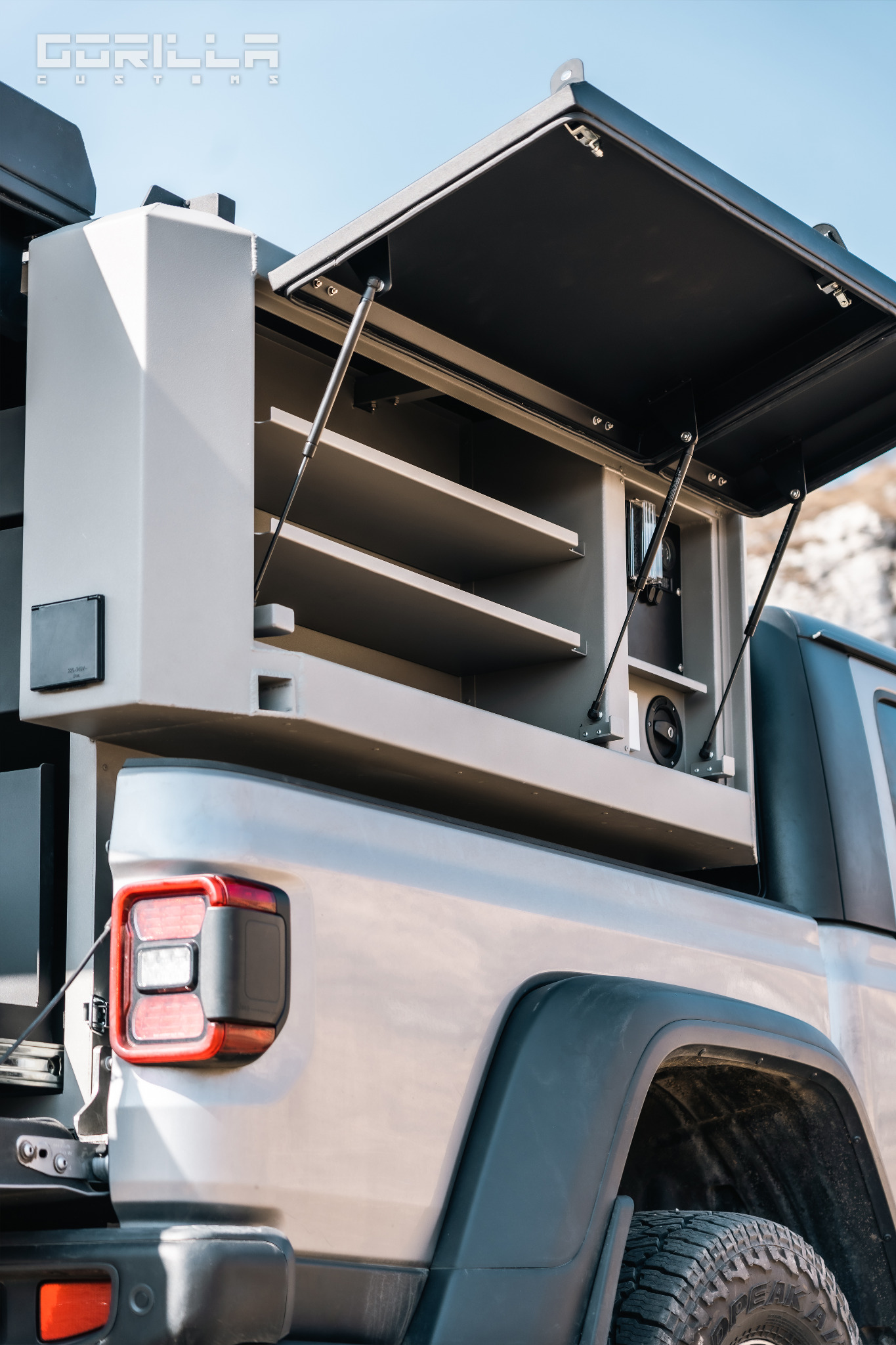 Szara zabudowa warsztatowa z półkami zamontowana na pace srebrnego pickupa, widok z tyłu, otwarte wieko. Widoczny tylny reflektor i opona terenowa.