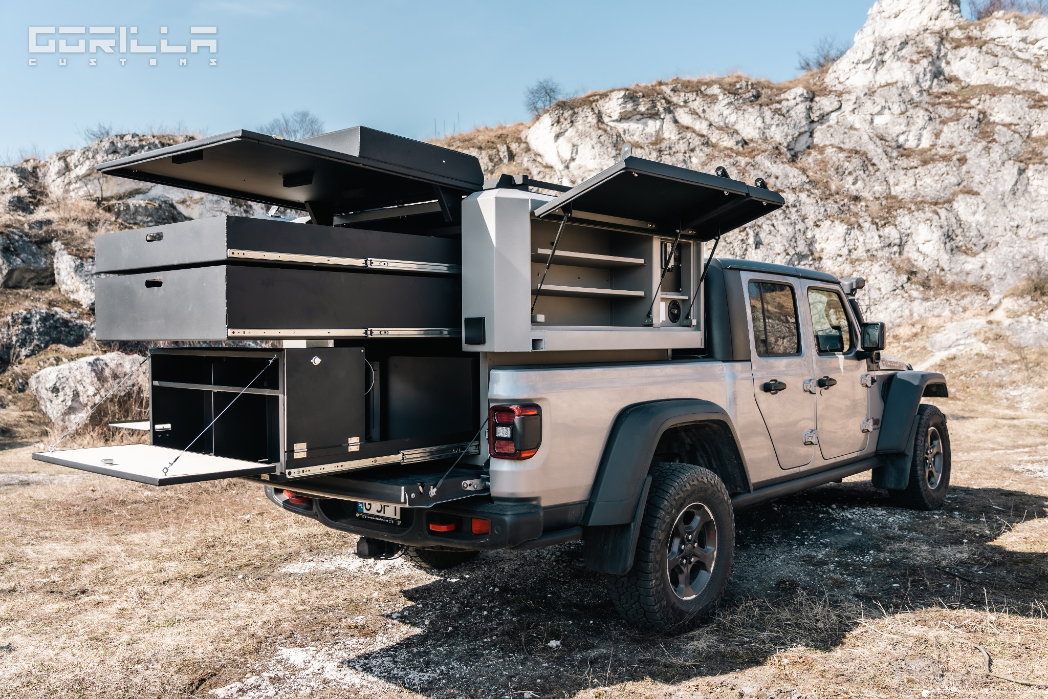 Szara terenówka z zabudową kempingową, otwarte szuflady i szafki, tło skaliste wzgórze.