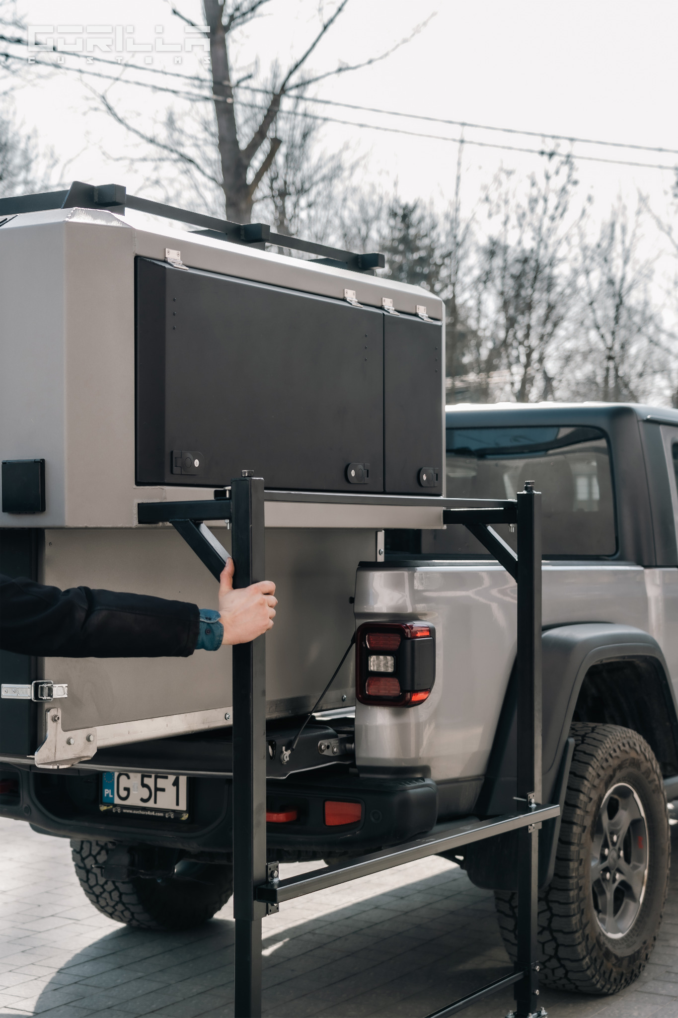 Szary pickup z zamontowaną zabudową kempingową, widoczne mocowania konstrukcji stalowej i dłoń w mankiecie koszuli.