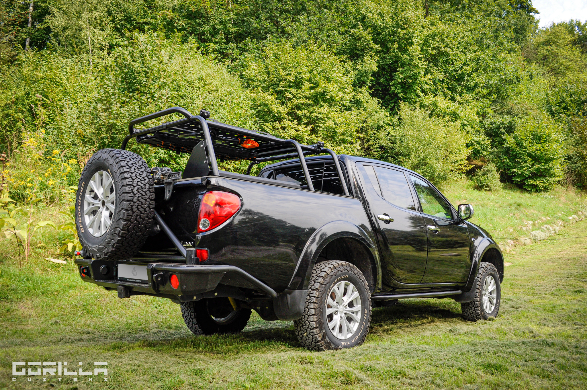 Czarny pickup z zamontowaną metalową konstrukcją bagażnika i kołem zapasowym na tylnym zderzaku, stojący na trawie w otoczeniu zieleni.