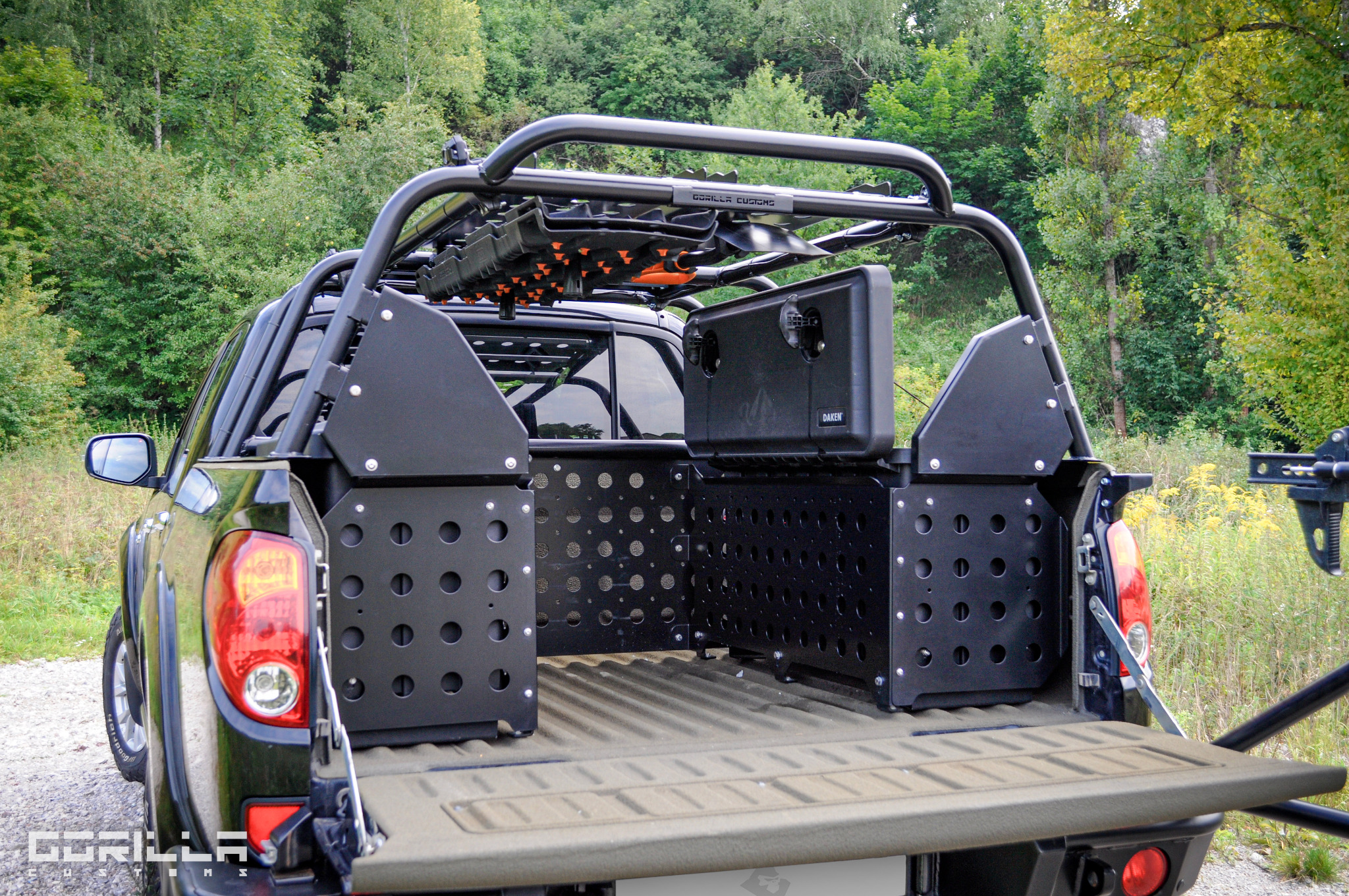 Czarny pickup z zamontowaną modułową zabudową przestrzeni ładunkowej z perforowanych paneli i dodatkowymi akcesoriami, takimi jak skrzynki narzędziowe i uchwyty na sprzęt terenowy, na tle zieleni.