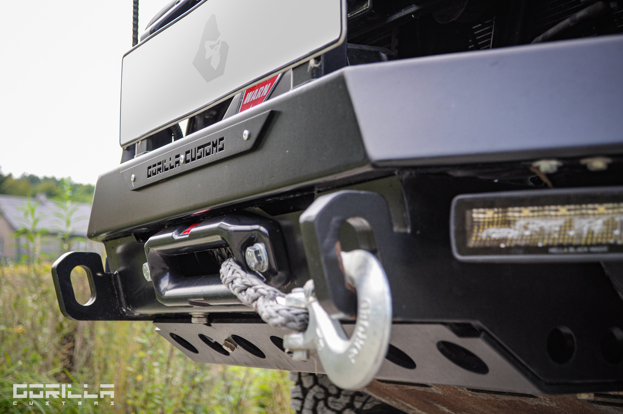 Czarny, stalowy zderzak terenowy z wyciągarką linową i uchwytami szekli, oświetleniem LED, logiem Gorilla Customs i tabliczką ostrzegawczą Warn.