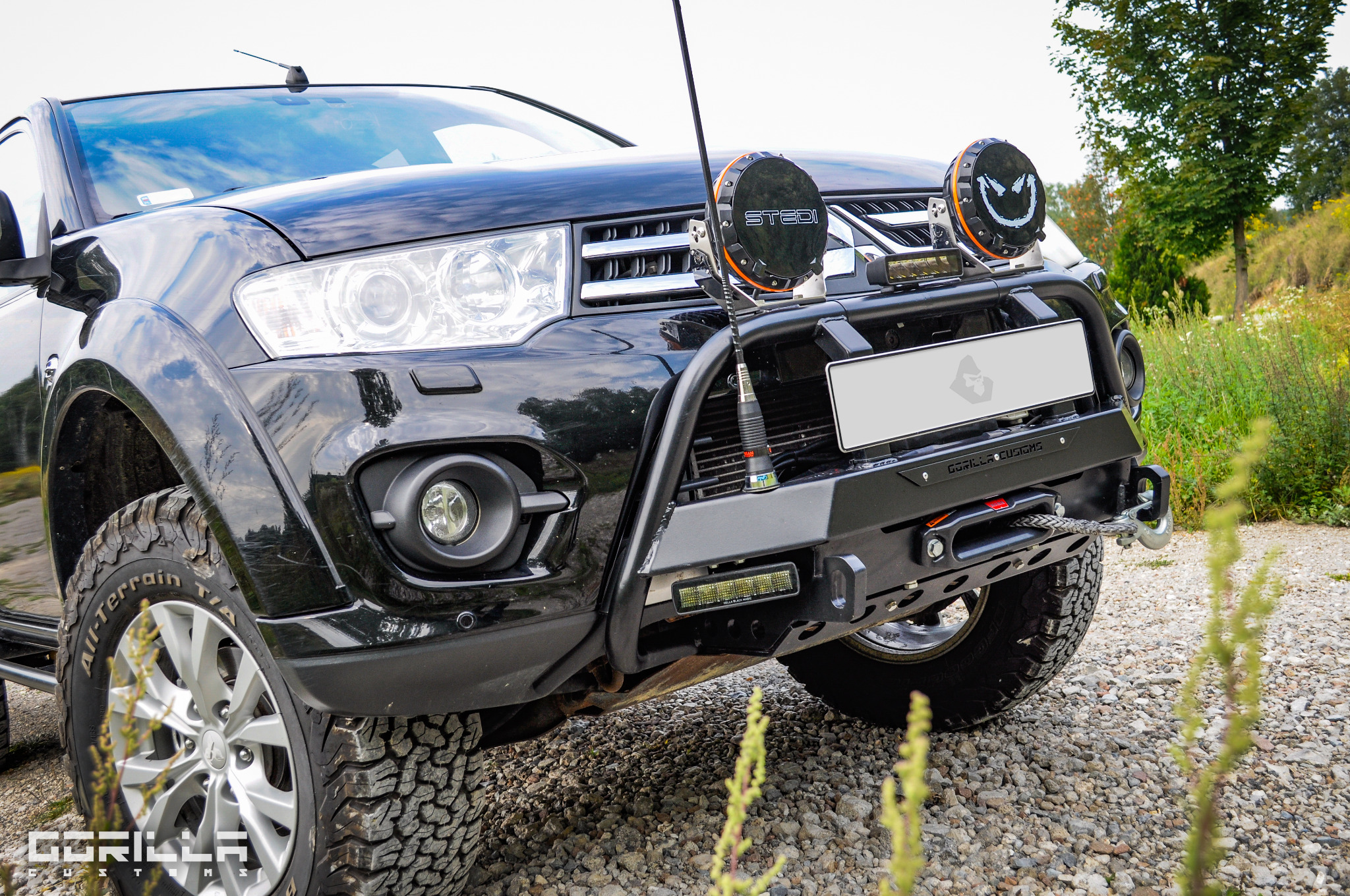 Czarny samochód terenowy z zamontowanym stalowym orurowaniem ochronnym, dodatkowym oświetleniem LED i wyciągarką linową, stojący na żwirowej drodze.