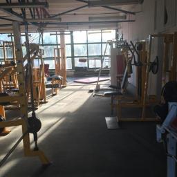 Old School GYM BY Marcin Kania - Przestronna siłownia z żółtymi maszynami do ćwiczeń, widok na okna z naturalnym światłem wpadającym do wnętrza, ciemna podłoga i surowa konstrukcja sufitu.