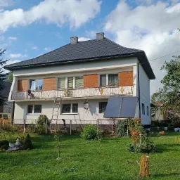 Dwupiętrowy dom z balkonem i panelami słonecznymi na elewacji, widok z ogrodu w słoneczny dzień.