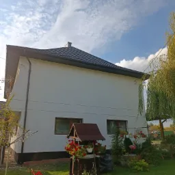 Ocieplona elewacja domu jednorodzinnego z czarnym dachem i elementami dekoracyjnymi w ogrodzie, widok z dołu w słoneczny dzień.