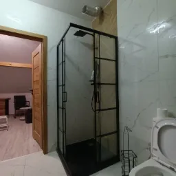 Nowoczesna łazienka z marmurowymi płytkami, czarną kabiną prysznicową typu walk-in i widocznym otwartym wejściem do innego pomieszczenia z drewnianymi elementami.