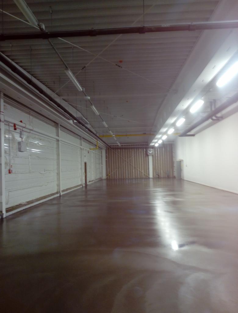 Błyszcząca, brązowa podłoga w hali przemysłowej z widocznymi liniami świetlnymi odbijającymi się od powierzchni, białe ściany i sufit z widocznymi instalacjami.