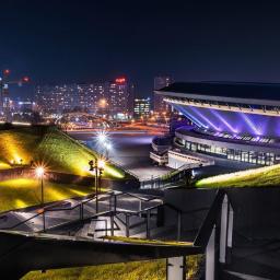 Fotografia Radosław Dybała - Nocny widok na Międzynarodowe Centrum Kongresowe w Katowicach, oświetlone fioletowymi reflektorami, z zielonymi wzgórzami i schodami w pierwszym planie oraz panoramą miasta w tle.