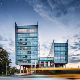 Fotografia Radosław Dybała - Nowoczesny budynek biurowy z przeszkloną fasadą, widoczny z poziomu ulicy z efektem rozmycia ruchu pojazdów, pod błękitnym niebem z chmurami.