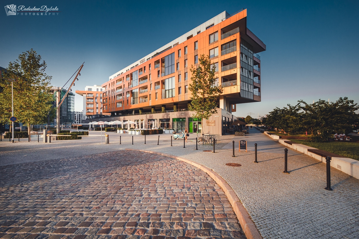 Nowoczesny budynek mieszkalny z pomarańczową elewacją, widoczny z poziomu brukowanej ulicy w słoneczny dzień.