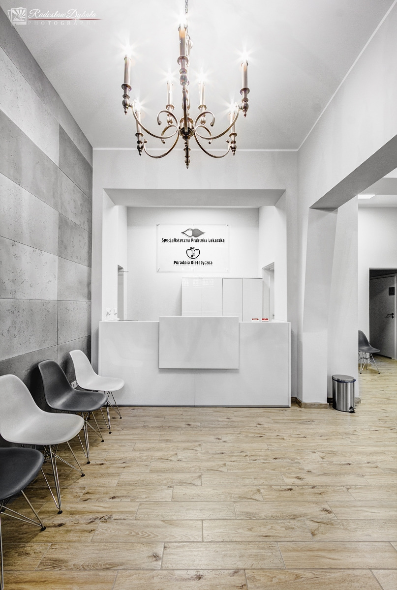 Jasne wnętrze poczekalni poradni dietetycznej z recepcją, wiszącym żyrandolem i rzędem krzeseł przy ścianie z betonowych paneli.