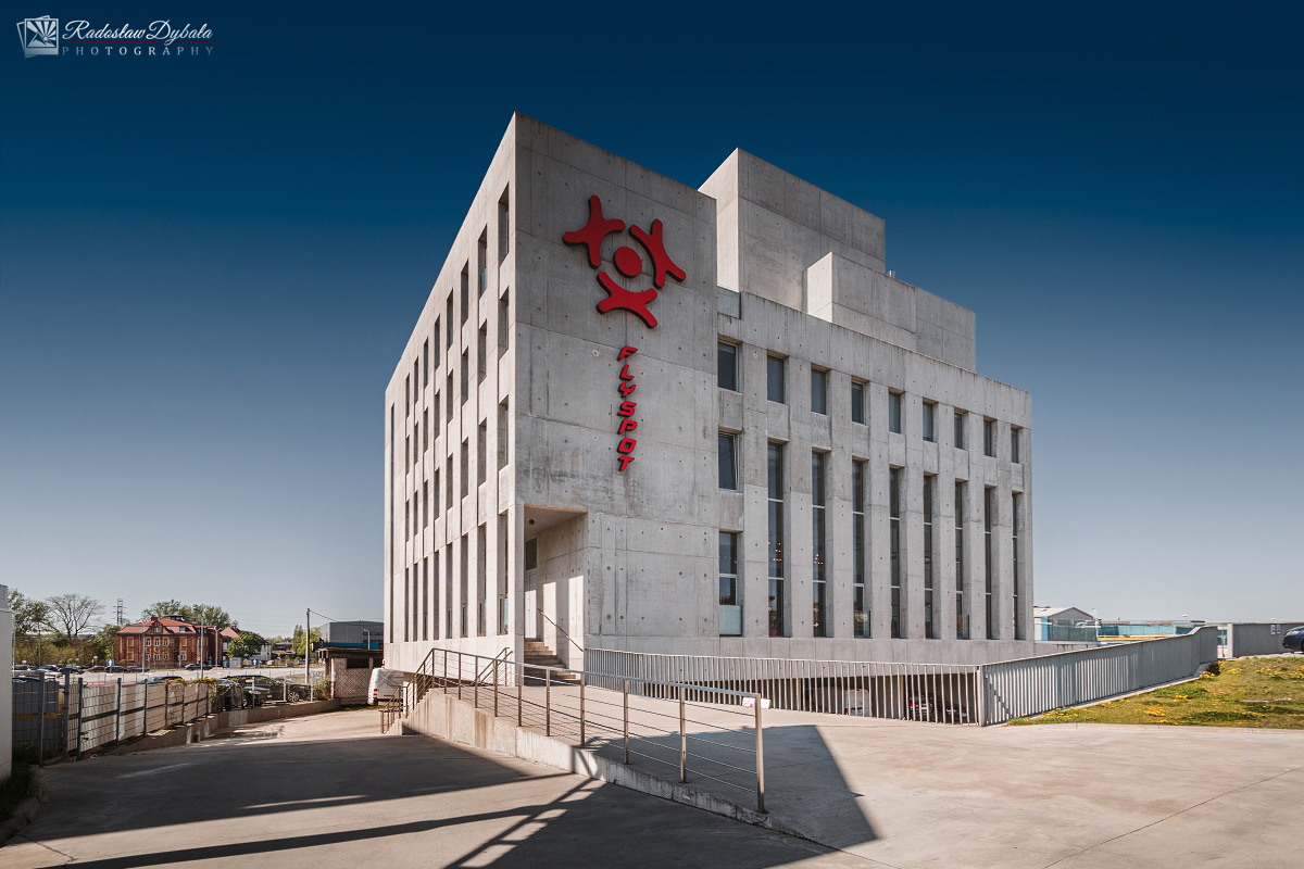 Betonowy budynek biurowy Flyspot z czerwonym logo i napisem, sfotografowany w słoneczny dzień z niskiej perspektywy.