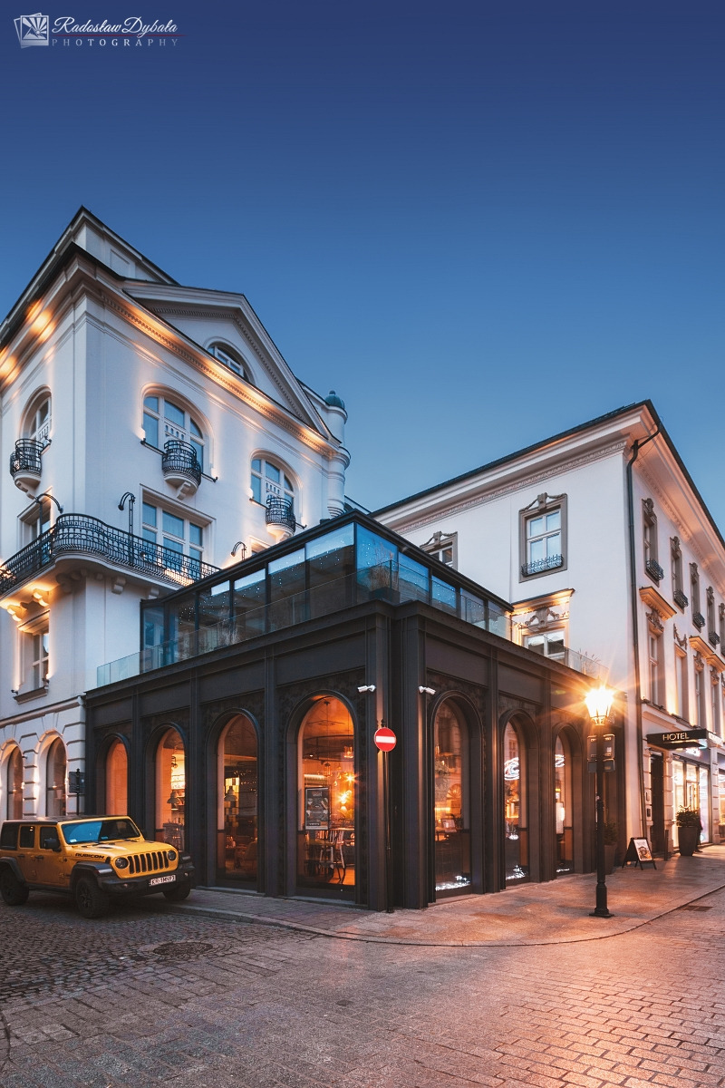 Elewacja kamienicy z przeszkloną dobudówką, widoczna żółta terenówka i oświetlenie uliczne