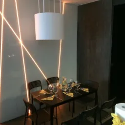 Minimalistyczna jadalnia z oświetleniem LED na ścianie, okrągłą lampą wiszącą i czarnym stołem nakrytym dla czterech osób z żółtymi serwetkami.