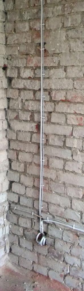 Pionowy przebieg przewodów elektrycznych mocowanych do surowej ściany z cegły za pomocą białych uchwytów, z widocznym fragmentem peszla przy podłodze.