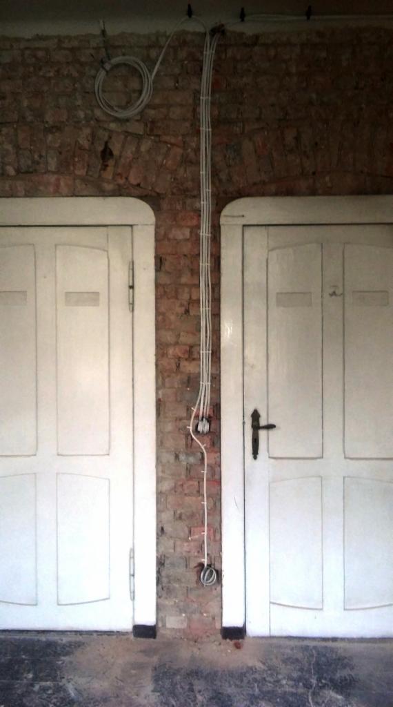 Kable elektryczne prowadzone po ceglanej ścianie między dwojgiem białych drzwi z panelami.