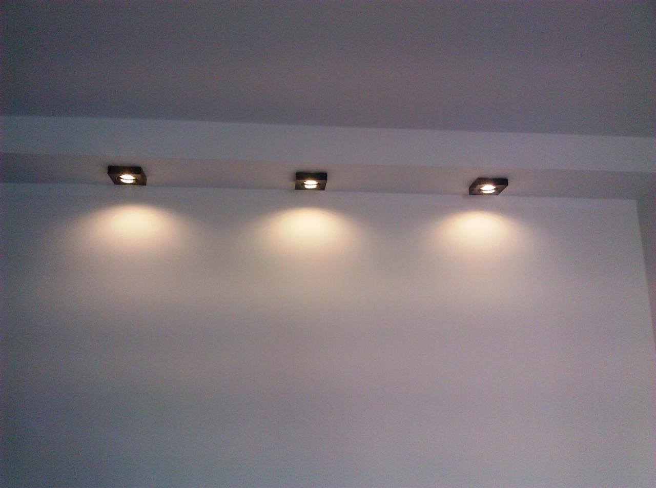 Trzy kwadratowe, czarne lampy sufitowe LED zamontowane w rzędzie na białej ścianie, emitujące ciepłe światło.