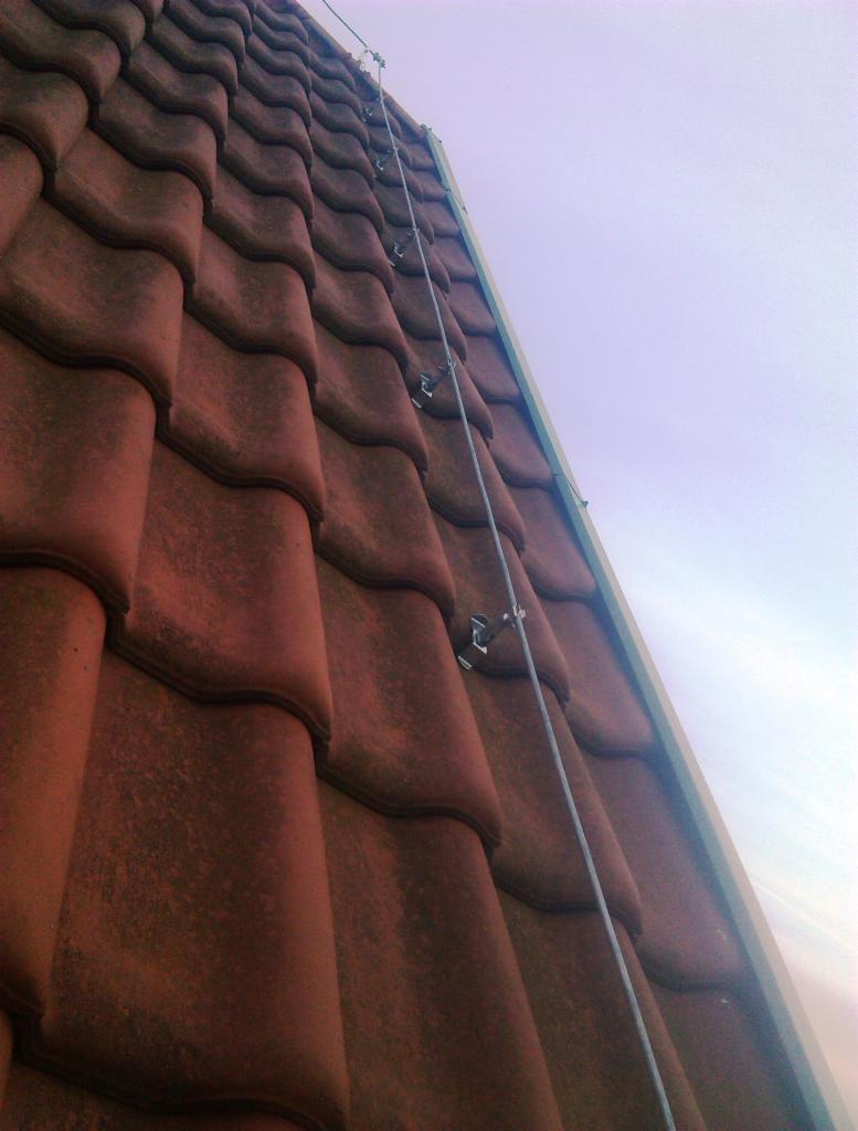 Czerwona dachówka ceramiczna z zamocowanym przewodem odgromowym i uchwytami, widok z dołu pod kątem w kierunku nieba.