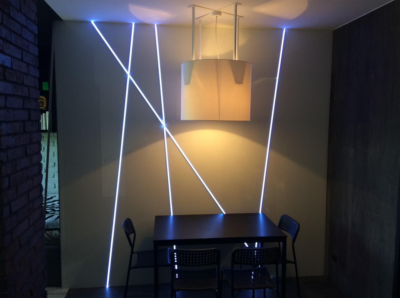 Jadalnia z nowoczesnym oświetleniem: lampa wisząca nad stołem i ukośne, błękitne linie LED na ścianie.