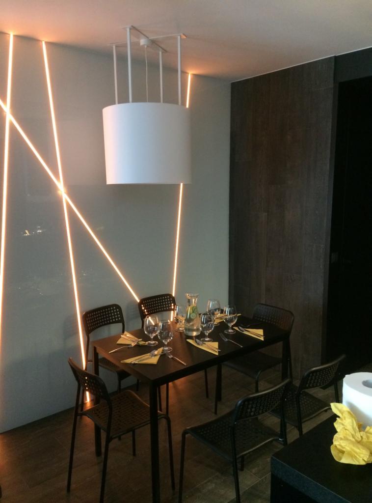Minimalistyczna jadalnia z oświetleniem LED na ścianie, okrągłą lampą wiszącą i czarnym stołem nakrytym dla czterech osób z żółtymi serwetkami.