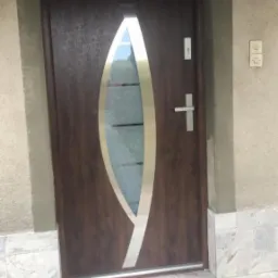 Brązowe drzwi zewnętrzne z owalnym przeszkleniem i srebrnymi akcentami, zamontowane w szarej ścianie, widoczne płytki marmurowe na podłodze.