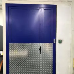 Dwuskrzydłowe, niebieskie drzwi z dolną częścią wykończoną blachą ryflowaną, zamontowane w surowym otworze z widocznymi śladami po pracach wykończeniowych, z czytnikiem kart dostępu po prawej stronie.