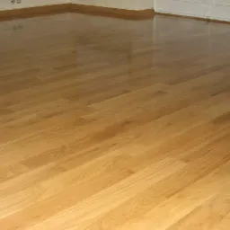 Błyszcząca, świeżo polakierowana podłoga z parkietu ułożonego w jodełkę, widoczne odbicia światła, jasne drewno, listwy przypodłogowe przy ścianie.