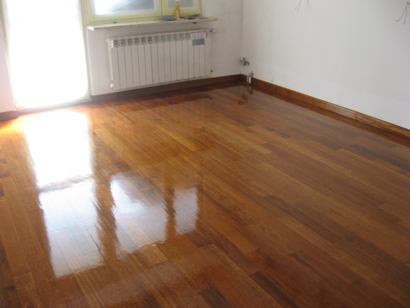 Błyszcząca, świeżo polakierowana podłoga z parkietu w odcieniach brązu, odbijająca światło z okna, widoczny biały grzejnik pod oknem i listwy przypodłogowe.