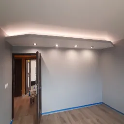 Szare wnętrze po odświeżeniu: pomalowane ściany, drewnopodobna podłoga, sufit podwieszany z oświetleniem punktowym i taśmą LED, otwarte drzwi wewnętrzne.