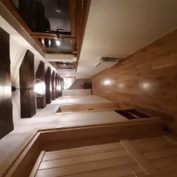 Wnętrze korytarza z drewnianą podłogą, białymi ścianami i dekoracyjnymi, ciemnymi belkami stropowymi oraz oknami dachowymi, oświetlone nowoczesnymi lampami.