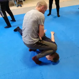 Instruktor demonstruje technikę obezwładniania na leżącym uczestniku kursu na niebieskiej macie, widoczne fragmenty innych osób w tle.