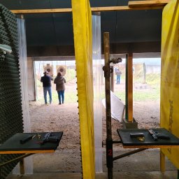 Szkoła Ochrony Kurs Ochrony STS Security Tactical Services Skierniewice - Strzelnica: osoby strzelają do tarcz, na stolikach broń i amunicja. Widok zza stanowiska, z żółtą przegrodą i numerem 2. Akustyczne panele na ścianach.
