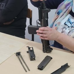Szkoła Ochrony Kurs Ochrony STS Security Tactical Services Skierniewice - Rozłożony pistolet maszynowy UZI na stole, widoczne elementy mechanizmu, ręce osoby składającej broń. Szkolenie z obsługi broni palnej.