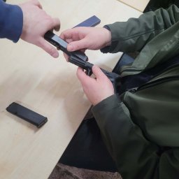 Szkoła Ochrony Kurs Ochrony STS Security Tactical Services Skierniewice - Instruktorzy prezentują rozkładanie i składanie broni palnej Glock na jasnym stole podczas kursu strzeleckiego. Widoczny magazynek i dłonie uczestników.