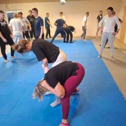 Szkoła Ochrony Kurs Ochrony STS Security Tactical Services Skierniewice - Sala treningowa z grupą osób ćwiczących techniki samoobrony na niebieskiej macie. Instruktor nadzoruje ćwiczenia. Widoczne lustra na ścianie.