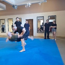 Szkoła Ochrony Kurs Ochrony STS Security Tactical Services Skierniewice - Trening sztuk walki na niebieskiej macie: mężczyzna w czarnej koszulce podnosi drugiego mężczyznę. W tle obserwatorzy i lustra.