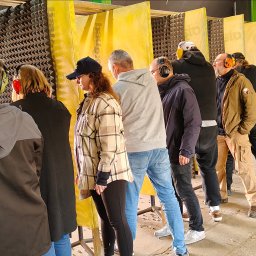 Szkoła Ochrony Kurs Ochrony STS Security Tactical Services Skierniewice - Grupa osób w słuchawkach ochronnych na strzelnicy, widoczne żółte przegrody i łuski nabojów na podłodze. Uczestnicy kursu strzeleckiego podczas zajęć praktycznych.